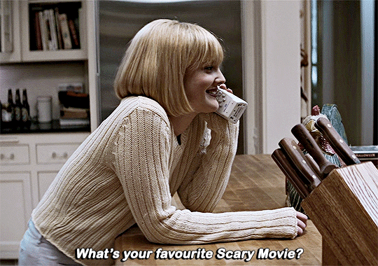 A GIF of a white woman with blonde bobbed hair, a cream sweater, and blue jeans, talking on a cordless landline telephone at a wooden kitchen counter. A block of knives is next to her.