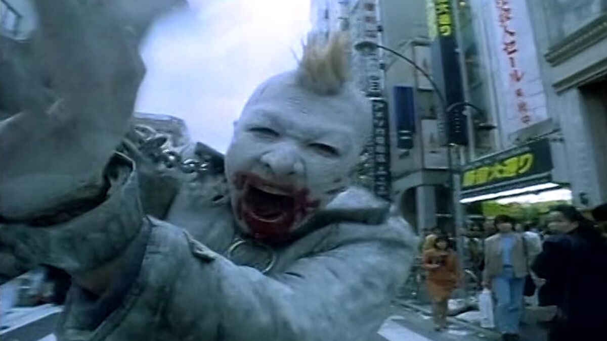 A still from the movie '964 Pinocchio'. A Japanese man is painted entirely white with what is likely blood smeared across his mouth. He has a blonde Mohawk and chains around his wrists as he screams at the camera.