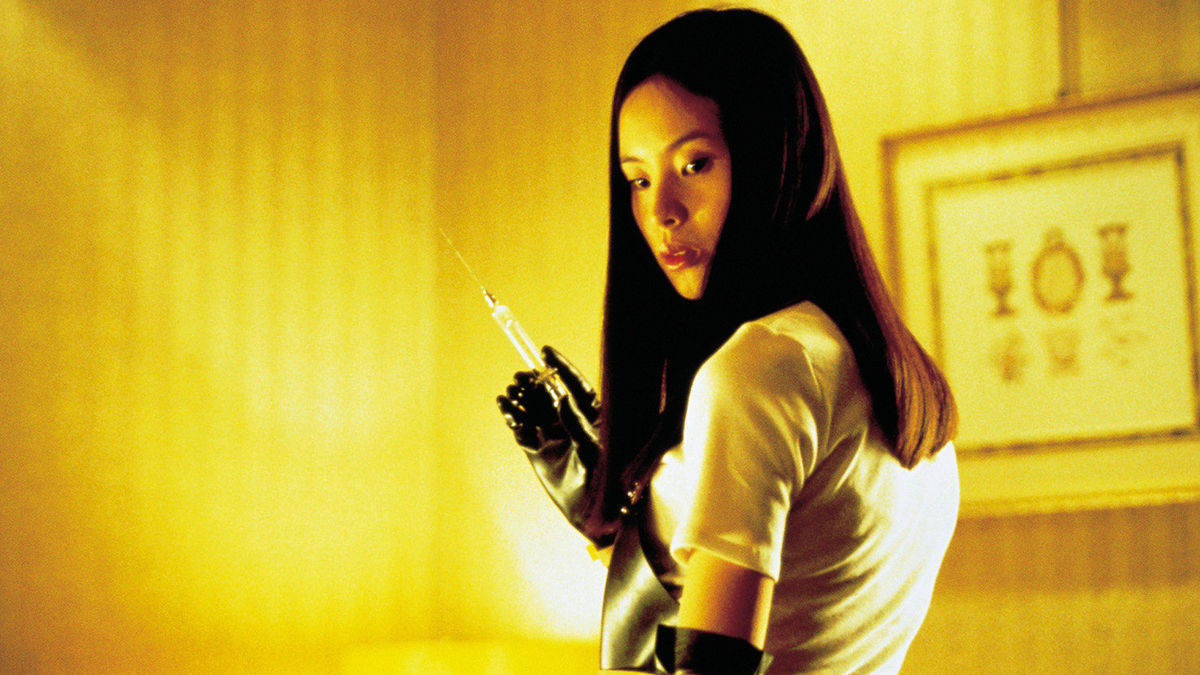 A still from the movie 'Audition. A Japanese woman with long hair holds a large syringe while wearing an apron and black gloves.