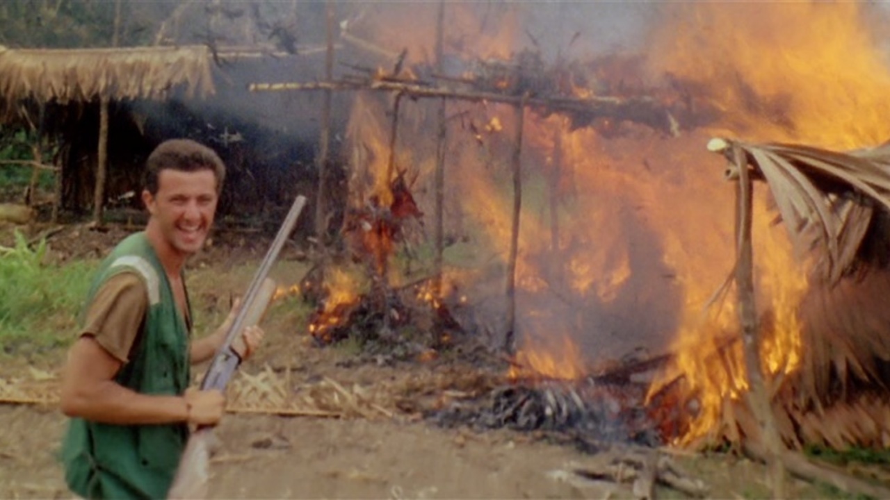 A still from 'Cannibal Holocaust'. A white man laughs while holding a shotgun. A burning hut is in the background.