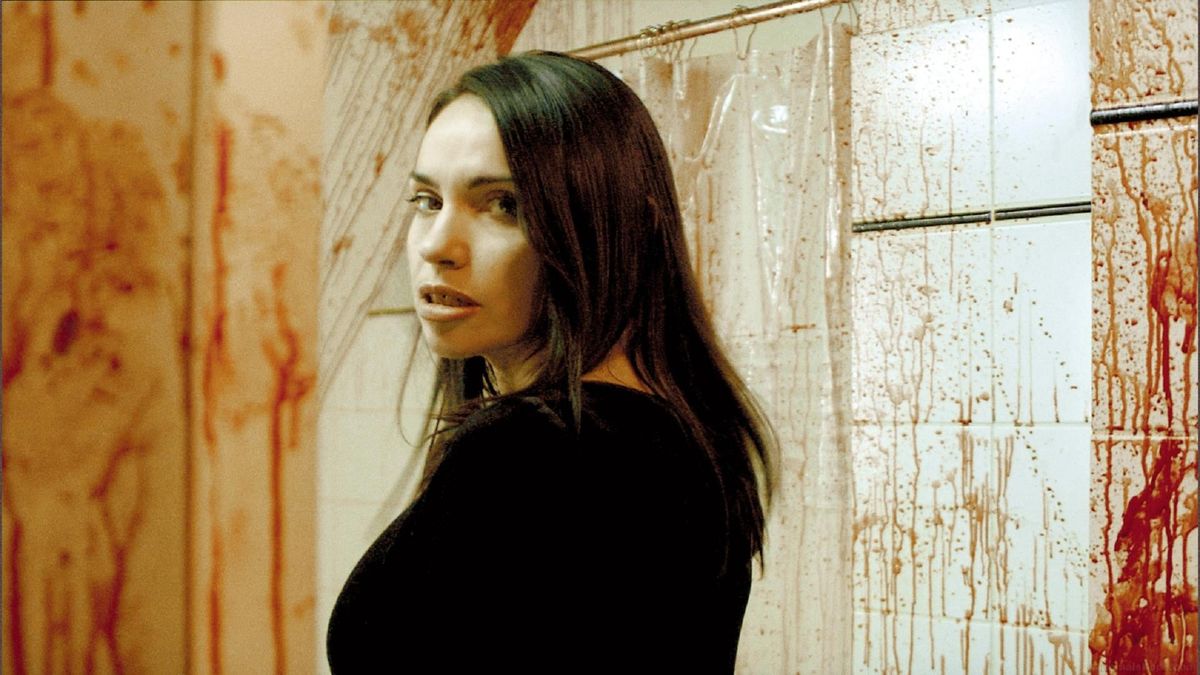 A still from the movie 'Inside'. A white woman with long brown hair stands in front of a white tiled wall that is splattered with blood.