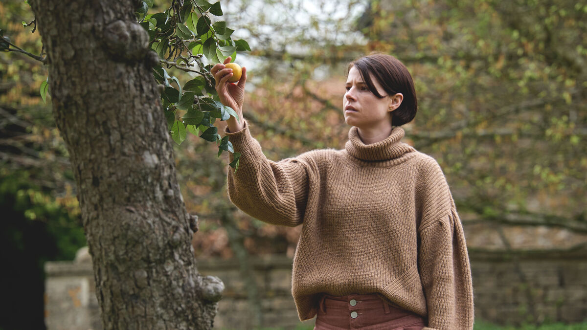 A still from the movie 'Men'. A white woman with a short brunette bob haircut picks fruit from a tree.