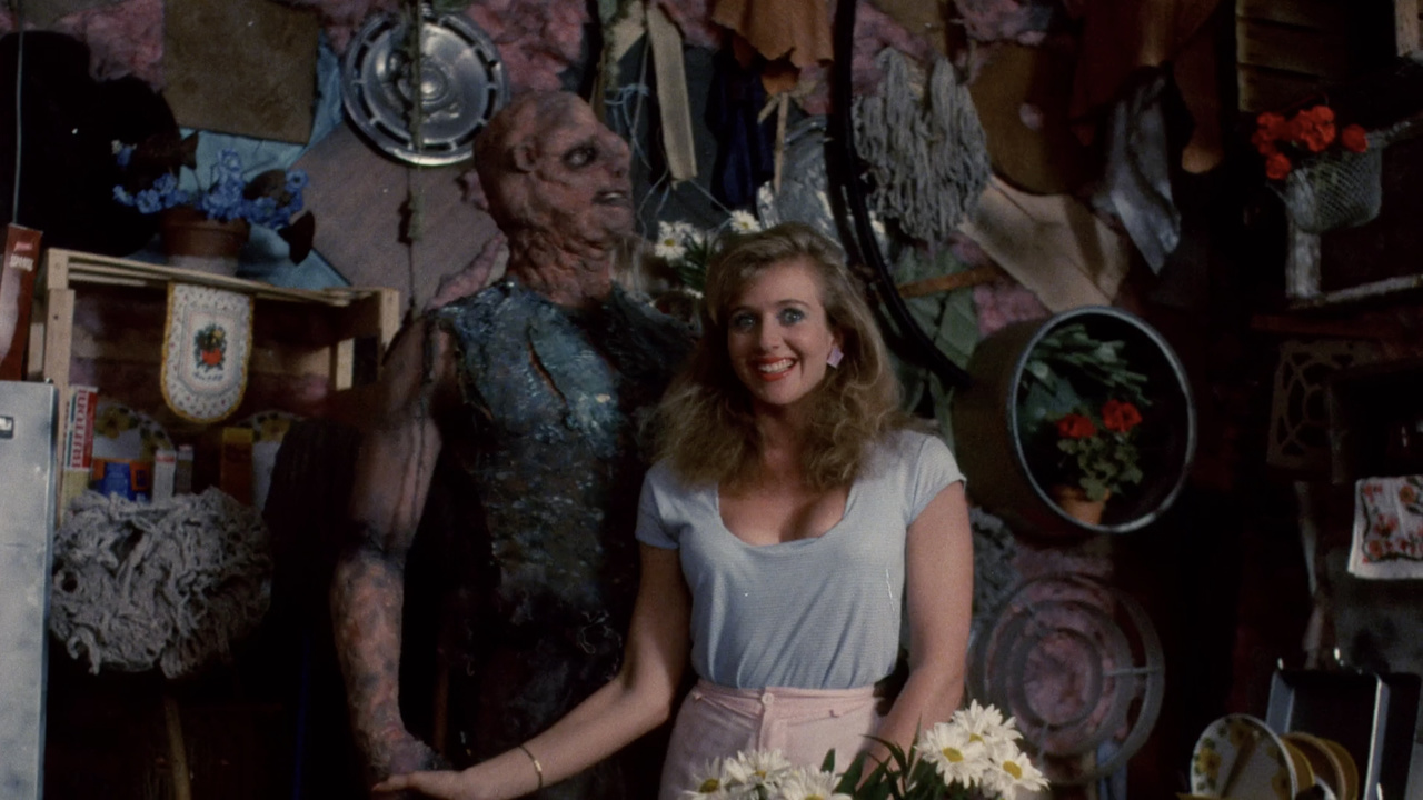 A still from 'The Toxic Avenger'. A tall humanoid monster stands next to a white woman who is smiling.